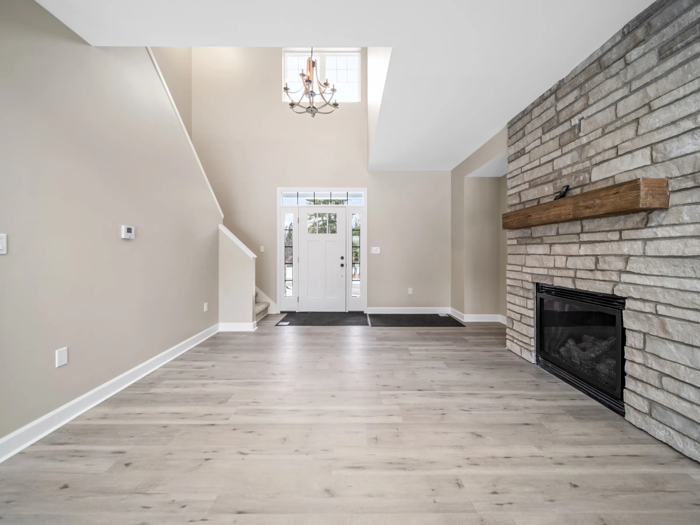 Two-story entry with a chandelier overhead, the front door flanked by glass, and the stone fireplace at the edge of the frame.