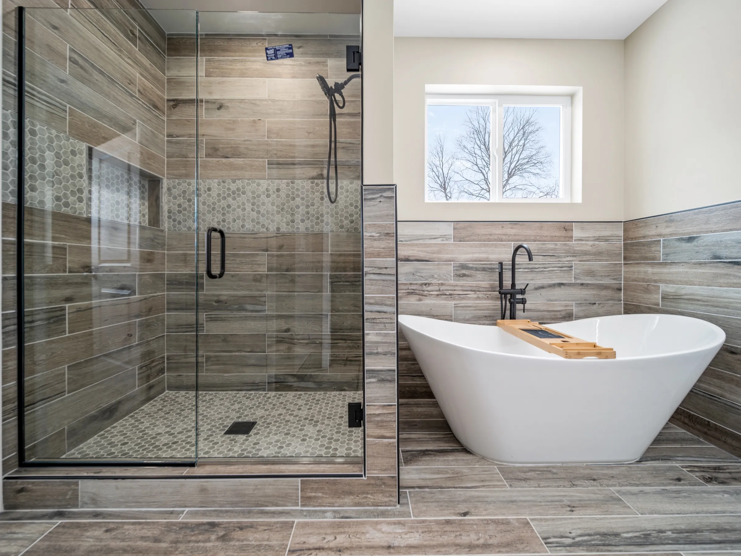 Primary bathroom with a freestanding soaking tub under the window, a black floor-mounted filler, and a glass shower tiled in wood-look plank.