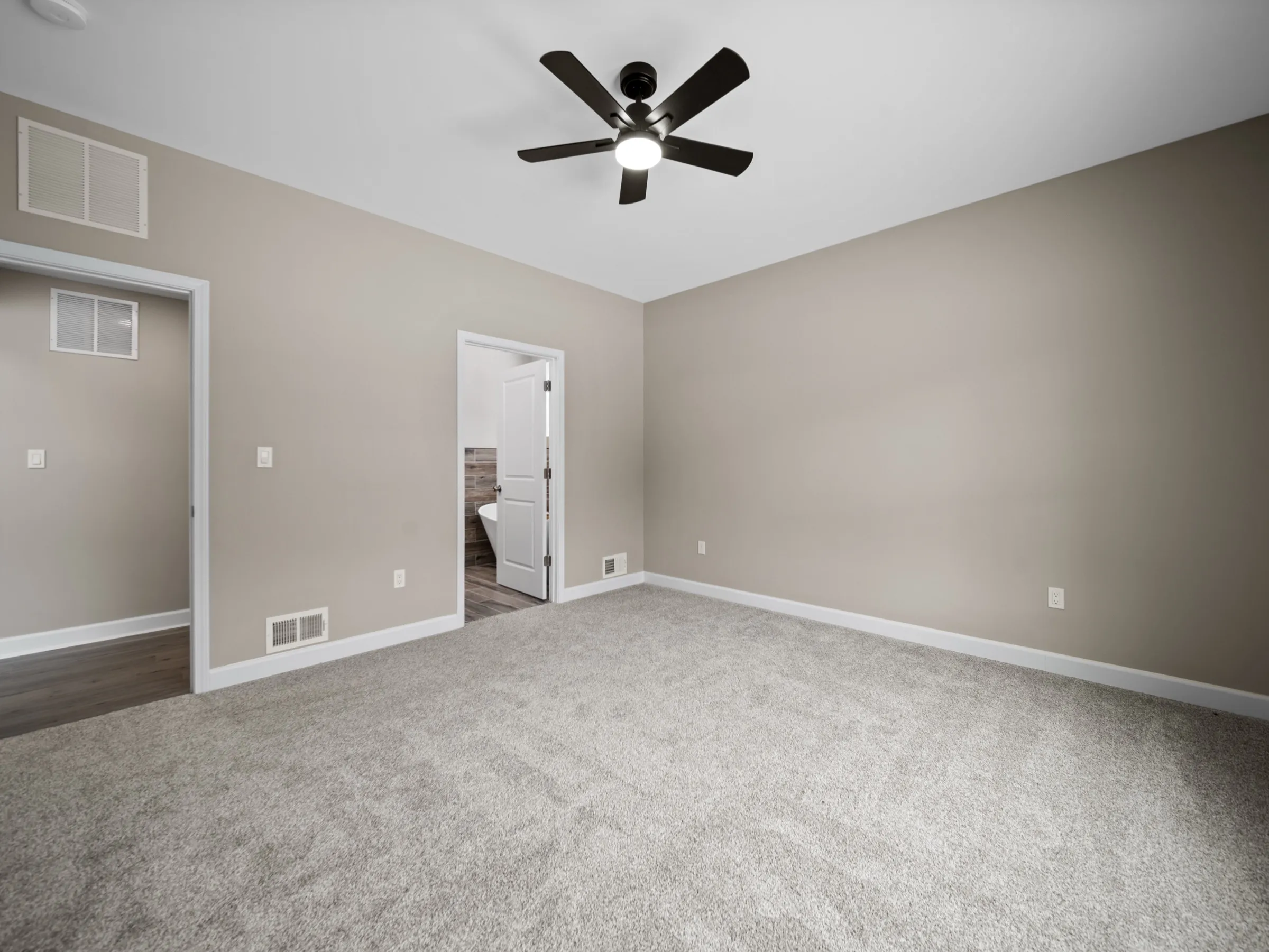 Carpeted primary bedroom with a ceiling fan, with the door open to the ensuite bath and the tub visible beyond.