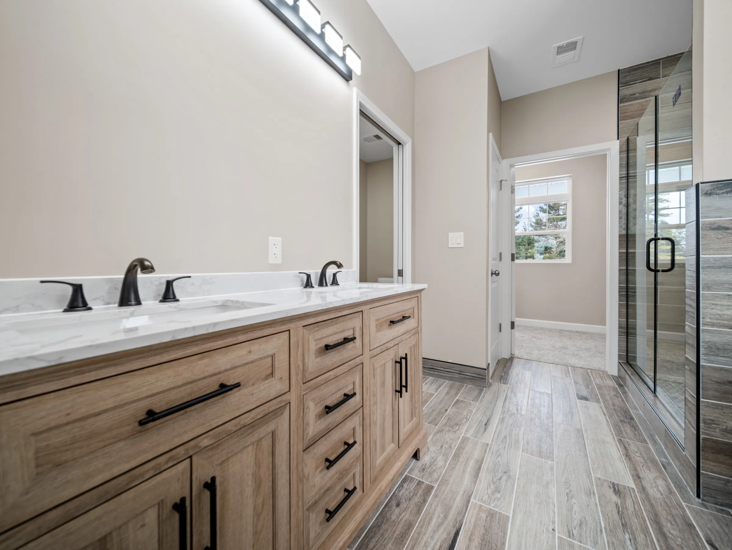 Double vanity in light oak with a marble-look top and black fixtures, with the glass shower reflected down the wood-look tile floor.