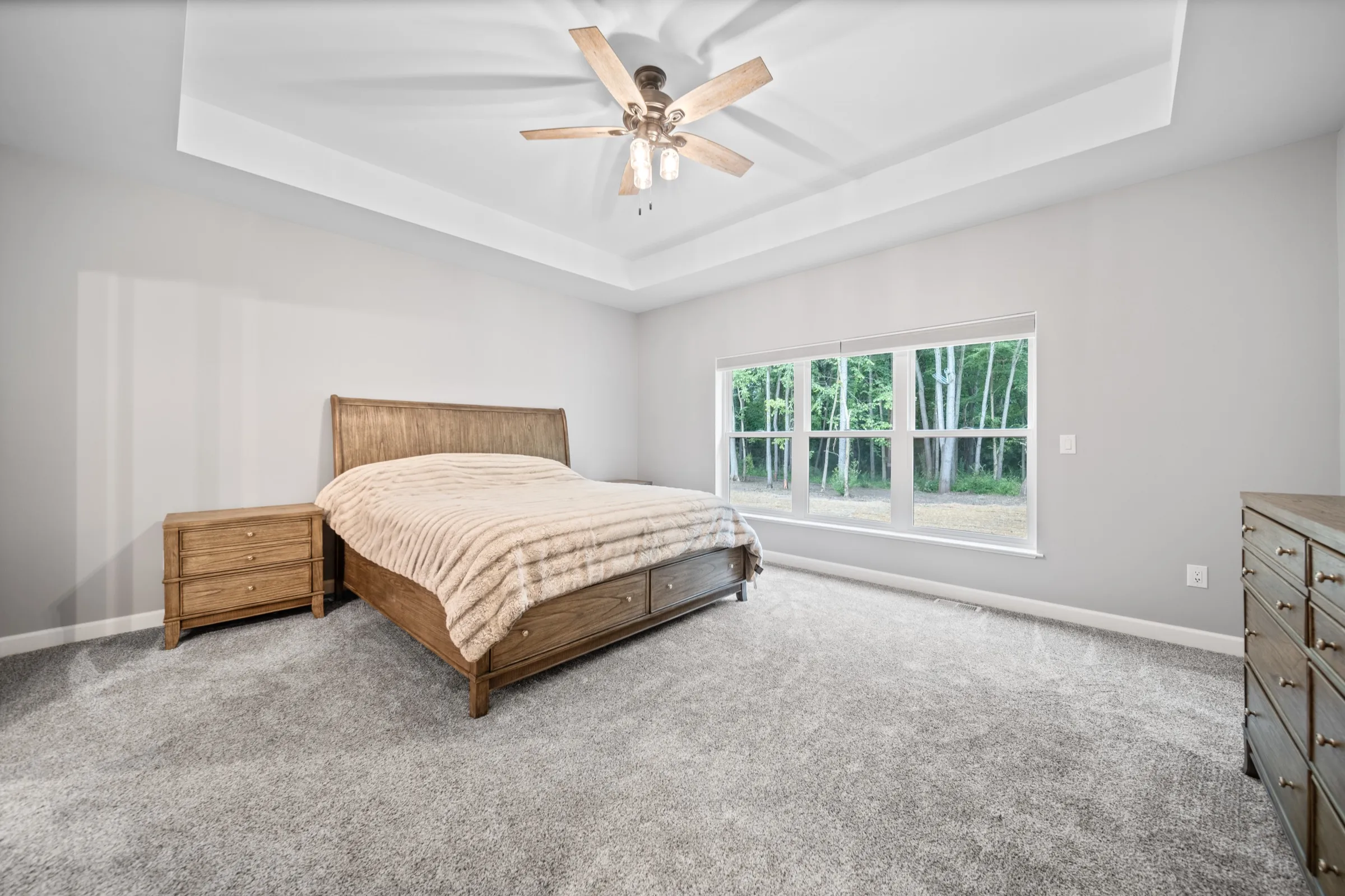 Primary bedroom with a tray ceiling, ceiling fan and windows onto the tree line.