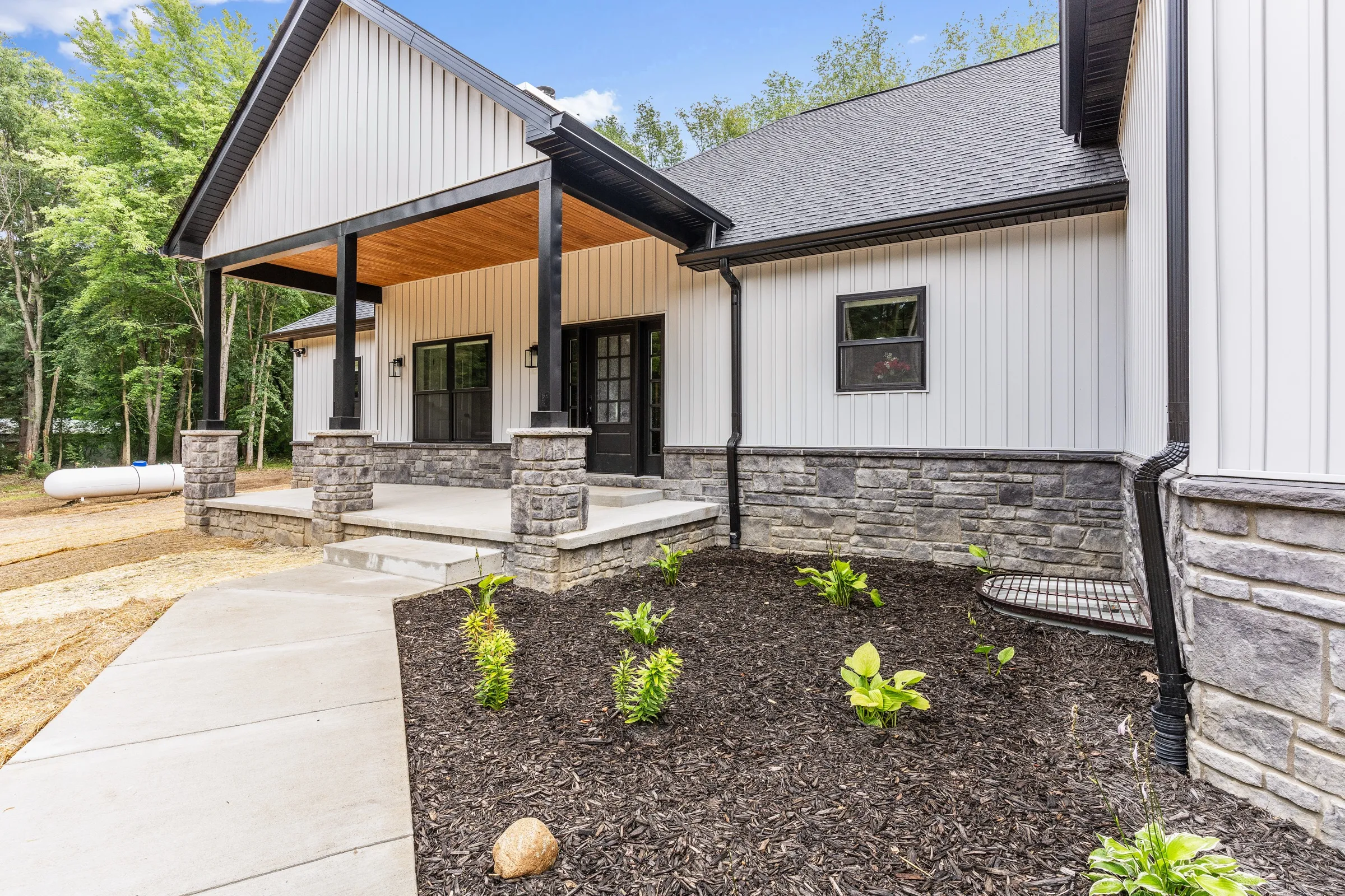 Front porch with stone-wrapped columns, black window frames, board and batten siding and a freshly planted bed.